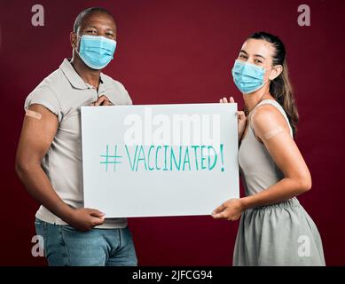 Covid a vacciné un homme afro-américain et une femme de race mixte montrant et tenant une affiche. Deux personnes portant un masque chirurgical isolé dans un studio rouge Banque D'Images