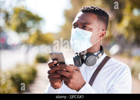 Homme noir méconnaissable portant un masque pour se protéger contre la pandémie du virus corona tout en portant des écouteurs et des SMS sur un téléphone portable Banque D'Images