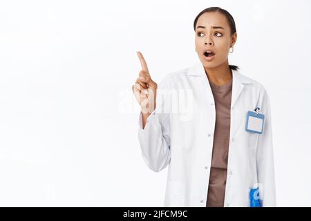 Portrait d'une femme médecin pointant, regardant le smth avec le visage concerné, choqué, se tient dans une couche blanche sur fond blanc Banque D'Images