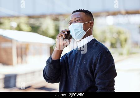 Homme d'affaires noir portant un masque et parlant sur un téléphone portable à l'extérieur. Faire un appel pendant les déplacements pendant la pandémie de covid Banque D'Images