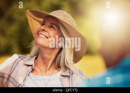 Femme aîée souriante appréciant une journée de dégustation de vin dans une ferme avec des amis. Bonne femme caucasienne portant un chapeau tout en étant assise et solidaire sur un vignoble Banque D'Images