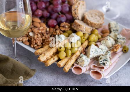 Verre de vin blanc et assiette avec différents en-cas.Fromage bleu, olives, tranches de baguette, graissini, jambon,raisins et noix.En-cas au vin sur le fond. Banque D'Images