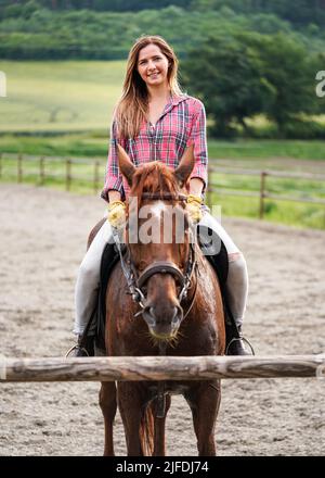 Jeune femme en chemise reposant sur un cheval brun - son manteau mouillé de la sueur - après l'équitation, souriant Banque D'Images