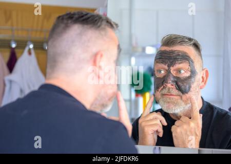 Homme barbu d'âge moyen utilisant un masque facial cosmétique vérifiant sa réflexion dans le miroir dans un concept de soins de la peau Banque D'Images