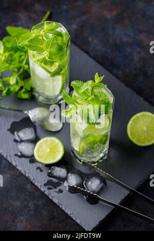 Cocktail Mojito, tranches de citron vert, menthe et glace sur fond sombre. Mojito à cocktail, pinces à glace et cuillère à cocktail sur plateau en ardoise. Vue de dessus Banque D'Images