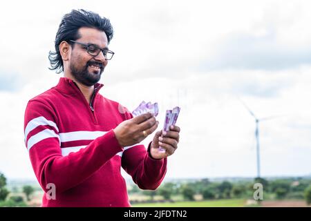 Homme souriant heureux comptant l'argent devant l'éolienne ou la centrale électrique - concept d'économie d'argent en utilisant l'énergie renouvelable et écologique Banque D'Images