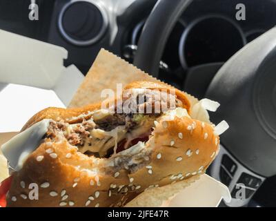 Manger en conduisant : un hamburger mordu dans les mains à l'intérieur de la voiture. Arrière-plan du volant. Banque D'Images