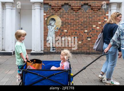 Famille passant la fresque d'art de rue par Hendog, un artiste anonyme, représentant une jeune fille cueillant des pétales d'une fleur, à Winchester, Hampshire, Royaume-Uni. Banque D'Images