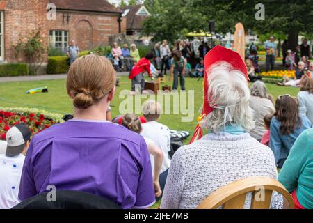 Travailleur de soins assis avec une dame âgée dans un parc qui regarde un amuseur clown, Angleterre, Royaume-Uni. Soignant aidant la personne âgée avec des activités sociales. Banque D'Images