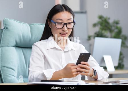 Jeune femme d'affaires asiatique travaille dans un bureau moderne, utilise le téléphone, sourit, lit les bonnes nouvelles, discute dans une application avec des amis Banque D'Images