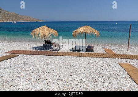 Les parasols et les chaises longues de plage de paille donnent sur une scène idyllique de plage avec ciel bleu, village de Livadia, île de Tilos, Dodcanese, Grèce Banque D'Images