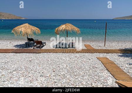 Les parasols et les chaises longues de plage de paille donnent sur une scène idyllique de plage avec ciel bleu, village de Livadia, île de Tilos, Dodcanese, Grèce Banque D'Images