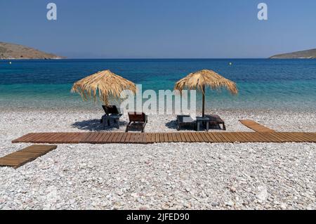 Les parasols et les chaises longues de plage de paille donnent sur une scène idyllique de plage avec ciel bleu, village de Livadia, île de Tilos, Dodcanese, Grèce Banque D'Images