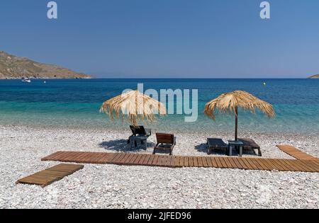 Les parasols et les chaises longues de plage de paille donnent sur une scène idyllique de plage avec ciel bleu, village de Livadia, île de Tilos, Dodcanese, Grèce Banque D'Images