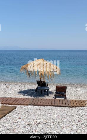 Les parasols et les chaises longues de plage de paille donnent sur une scène idyllique de plage avec ciel bleu, village de Livadia, île de Tilos, Dodcanese, Grèce Banque D'Images