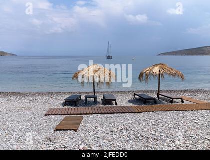 Les parasols et les chaises longues de plage de paille donnent sur une scène idyllique de plage avec ciel bleu, village de Livadia, île de Tilos, Dodcanese, Grèce Banque D'Images