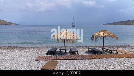 Les parasols et les chaises longues de plage de paille donnent sur une scène idyllique de plage avec ciel bleu, village de Livadia, île de Tilos, Dodcanese, Grèce Banque D'Images