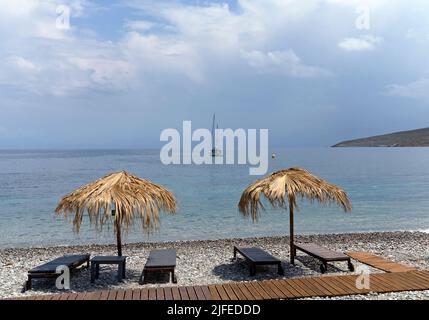 Les parasols et les chaises longues de plage de paille donnent sur une scène idyllique de plage avec ciel bleu, village de Livadia, île de Tilos, Dodcanese, Grèce Banque D'Images