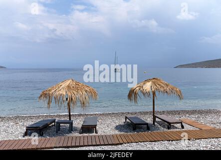 Les parasols et les chaises longues de plage de paille donnent sur une scène idyllique de plage avec ciel bleu, village de Livadia, île de Tilos, Dodcanese, Grèce Banque D'Images