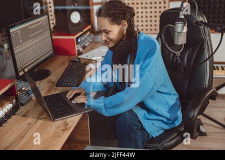 Joyeuse femme africaine américaine compositeur, producteur, musicienne, anchorwoman tapant du texte sur ordinateur portable tout en travaillant dans la salle de l'ingénieur du son dans les profes modernes Banque D'Images