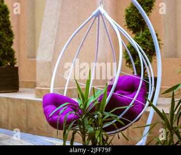 Chaise à bascule en rotin extérieure contre un mur orange. Chaise en osier blanche et confortable avec coussin tapissé de violet dans un coin salon au milieu de la tropica Banque D'Images