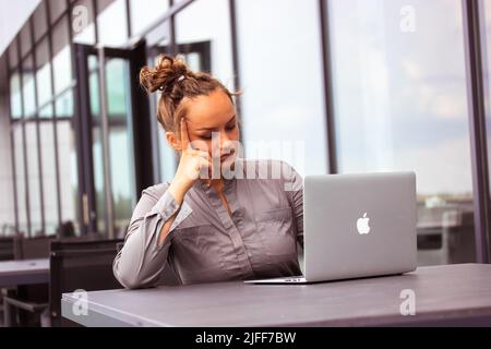 Kiev, Ukraine, 17 juillet 2021. Femme d'affaires sérieuse utilisant l'ordinateur portable Apple MacBook assis sur la terrasse du café. Latina fille en costume gris travaille à distance. Banque D'Images