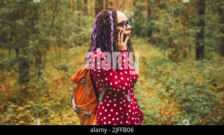 Une jeune femme perdue dans des lunettes avec sac à dos parlant sur un téléphone portable, debout dans la forêt estivale. Touriste féminin parlant sur smartphone, errant dans les bois Banque D'Images