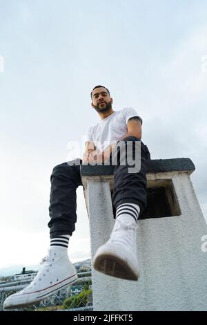 portrait de jeune homme modèle avec la barbe et les cheveux courts assis posant dans le t-shirt blanc, jeans et chaussures de sport, l'homme latin attrayant Banque D'Images