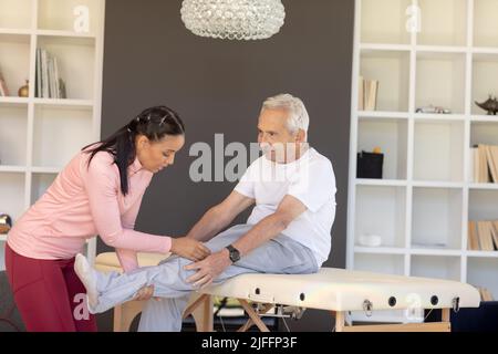 Kinésithérapeute biracial féminin donnant un massage de jambe à l'homme âgé caucasien à la maison Banque D'Images