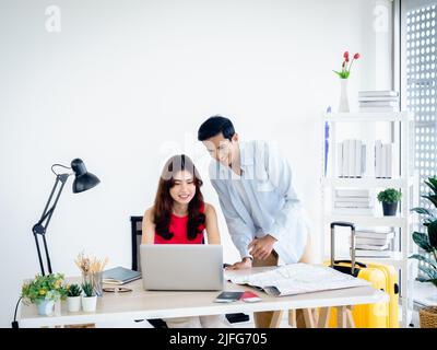 Joyeux couple asiatique, jeune femme et homme utilisant ordinateur portable et carte ensemble pour la réservation de vol, la chambre d'hôtel et les informations de voyage dans le bureau, prêt à Banque D'Images