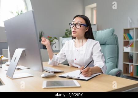 Jeune belle asiatique médecin consulte les patients consultation en ligne à distance, femme travaille dans le bureau utilise l'ordinateur et la webcam pour les appels vidéo. Banque D'Images