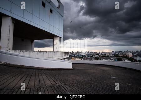 Vue en perspective d'un parking vide sur le toit d'un centre commercial abandonné, des nuages de pluie sombres couvrent une ville en arrière-plan. Banque D'Images