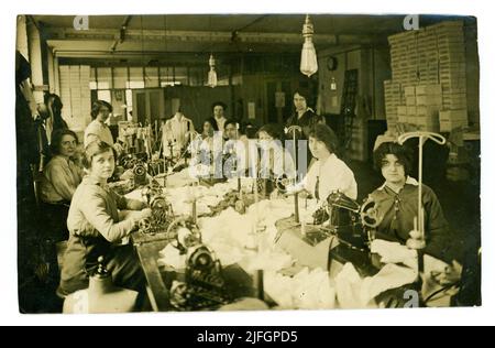 Photo originale de WW1 ans d'un groupe de jeunes femmes machinistes de classe ouvrière travaillant dans une usine de fabrication de chemisiers à l'aide de machines à coudre à pédales, un chemisier est exposé sur un mannequin de couturière, chaque dame a un stand de comptoir de couturière pour mettre le chemisier fini sur. Une direction regarde en arrière-plan. Les premières ampoules sont suspendues au-dessus de l'établi. Vers 1916. ROYAUME-UNI Banque D'Images