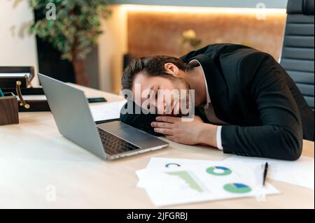 Exténué homme d'affaires caucasien fatigué, pdg, directeur d'entreprise, dans un costume formel, dormant sur le lieu de travail après une dure journée de travail, employé de bureau a besoin de repos, souffre de fatigue chronique et manque de sommeil Banque D'Images