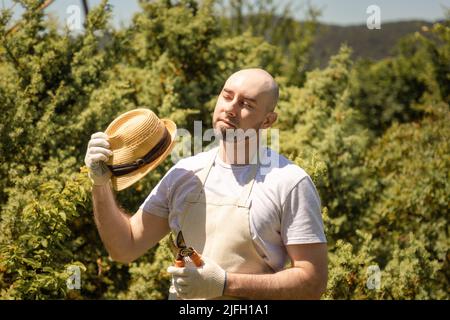 Coup de soleil. Portrait d'un homme à barbe chauve portant des gants et un tablier avec un chapeau de paille tenant un piston. En arrière-plan se trouve un arbre de jardin ensoleillé Banque D'Images