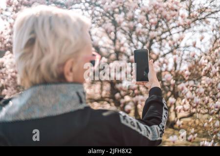 Beauté Caucasien 50s femme prendre des photos rose délicat fleur magnolia par le smartphone avec fond de fleur de foyer doux. Saison de printemps avec Banque D'Images