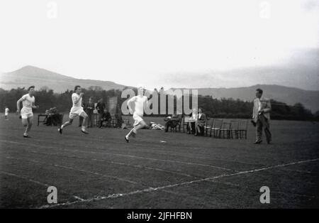 1952, historique, à l'extérieur d'un champ avec des montagnes au loin, trois garçons d'école senior se disputant une course de sprint sur une piste d'herbe, en Écosse, au Royaume-Uni, avec d'autres écoliers et deux enseignantes assis sur des chaises. Banque D'Images