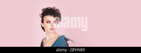 Une fille avec un grand sourire sincère se tient dans le studio sur un fond rose pastel. Une fille avec une coiffure étrange volant et maquillage brillant. Web Banque D'Images