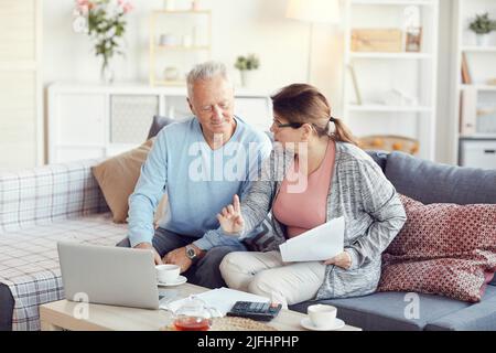 Un couple âgé sérieux et occupé vêtait de vêtements décontractés, assis sur un canapé dans la salle de séjour et calculant les factures tout en discutant du budget familial Banque D'Images