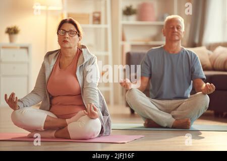 Couple sénior détendu et serein avec des yeux fermés assis avec des jambes croisées au sol dans le salon et pratiquant la méditation Banque D'Images