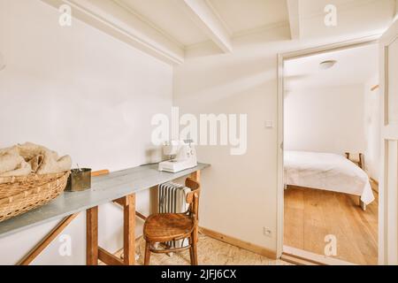 Une table avec machine à coudre, un panier, une chaise et un radiateur dans une pièce lumineuse avec un plafond à poutres apparentes blanches, à côté de la chambre Banque D'Images
