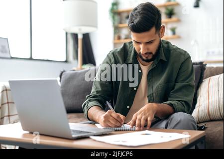 Spécialiste indien ou arabe, étudiant ou indépendant, assis à la maison sur un canapé dans la salle de séjour, utilisant un ordinateur portable, travaillant ou étudiant à distance, écoutant un webinaire, prenant des notes, faire des devoirs Banque D'Images