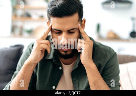 Maux de tête, spasme. Jeune homme de nationalité arabe ou indienne triste épuisé, assis sur le canapé dans le salon, movie ses temples, fermé ses yeux, se sent mal, migraine, fatigue, nécessite un traitement Banque D'Images