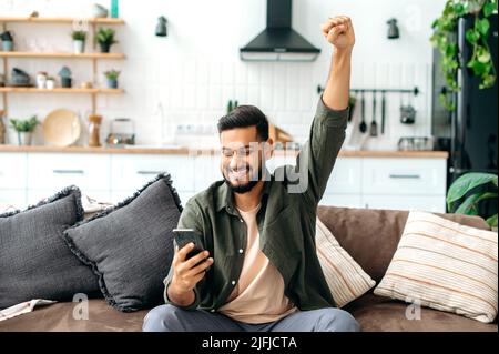 Gagner, succès. Indien joyeux jeune indien ou arabe en tenue décontractée, asseyez-vous à la maison dans le salon sur un canapé confortable, en utilisant le téléphone intelligent, lisez les bonnes nouvelles, en faisant des gestes à la main, souriant joyeusement Banque D'Images