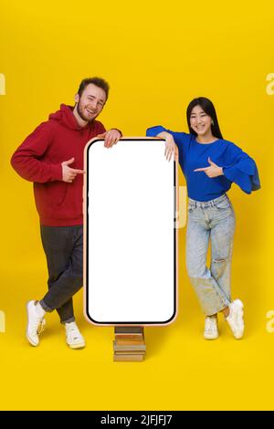 Un homme caucasien et une fille asiatique se pencha sur un immense smartphone géant, debout sur de vieux livres avec un écran blanc pointant sur lui regardant la publicité d'application mobile d'appareil photo isolée sur fond jaune. Banque D'Images