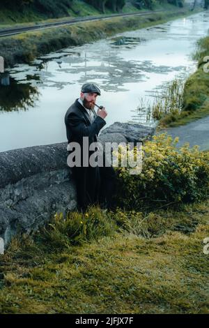 Gangster élégant homme assis à côté du canal irlandais et des voies de chemin de fer, fumer un tuyau au coucher du soleil. thème 1920s. Homme à la mode, brutal et confiant, barbu. Banque D'Images