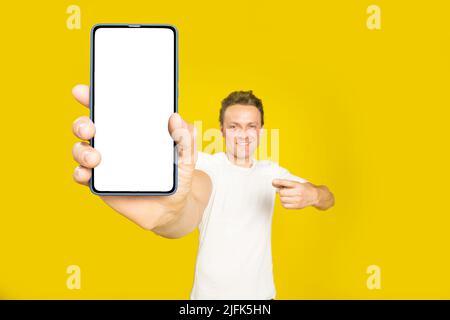 Application mobile publicitaire beau blond homme pointant vers un smartphone géant avec écran blanc vide dans sa main, portant un t-shirt blanc et un Jean isolé sur fond jaune. Offre exceptionnelle. Banque D'Images