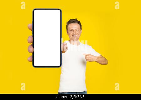 Beau homme blond pointant vers un smartphone géant avec écran blanc vide, portant un t-shirt blanc et un Jean écran de téléphone portable maquette isolée sur fond jaune. Publicité pour applications mobiles. Banque D'Images