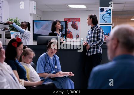 Patient asiatique parlant avec le réceptionniste de l'hôpital debout à la réception pendant que l'infirmière vérifie une jeune fille dans la salle d'attente. Jeune homme assistant à un rendez-vous de médecin en clinique avec divers patients. Banque D'Images