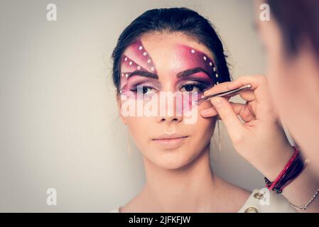 Après avoir peint son visage avec du violet, l'artiste de maquillage applique des cristaux sur le visage du jeune modèle famale avec des pinces Banque D'Images